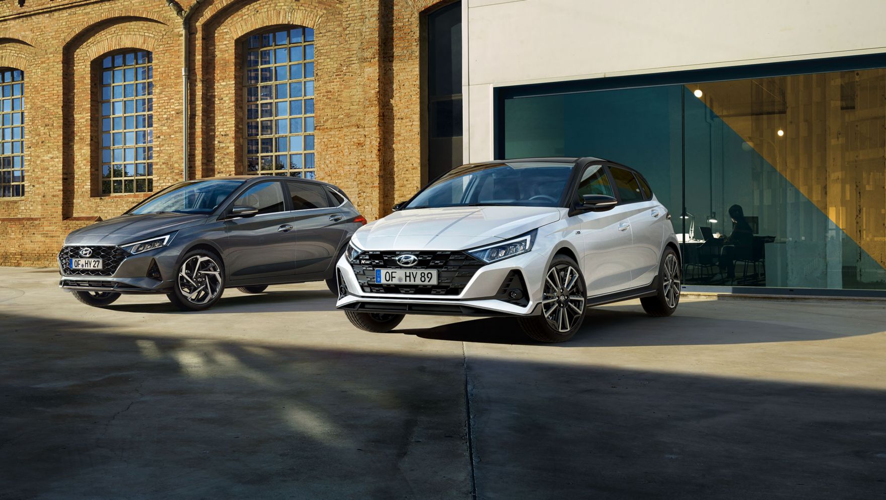 Nové vozy Hyundai i20 N Line a i20 v barvě aurora grey, zaparkované před velkou budovou.